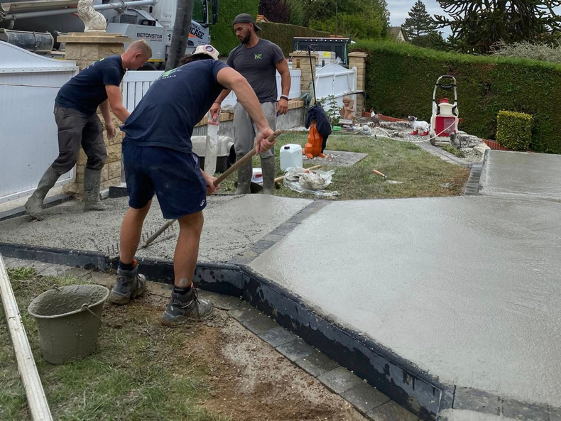 Trois ouvriers du bâtiment en chemises sombres lissent le béton frais d'une allée résidentielle.
