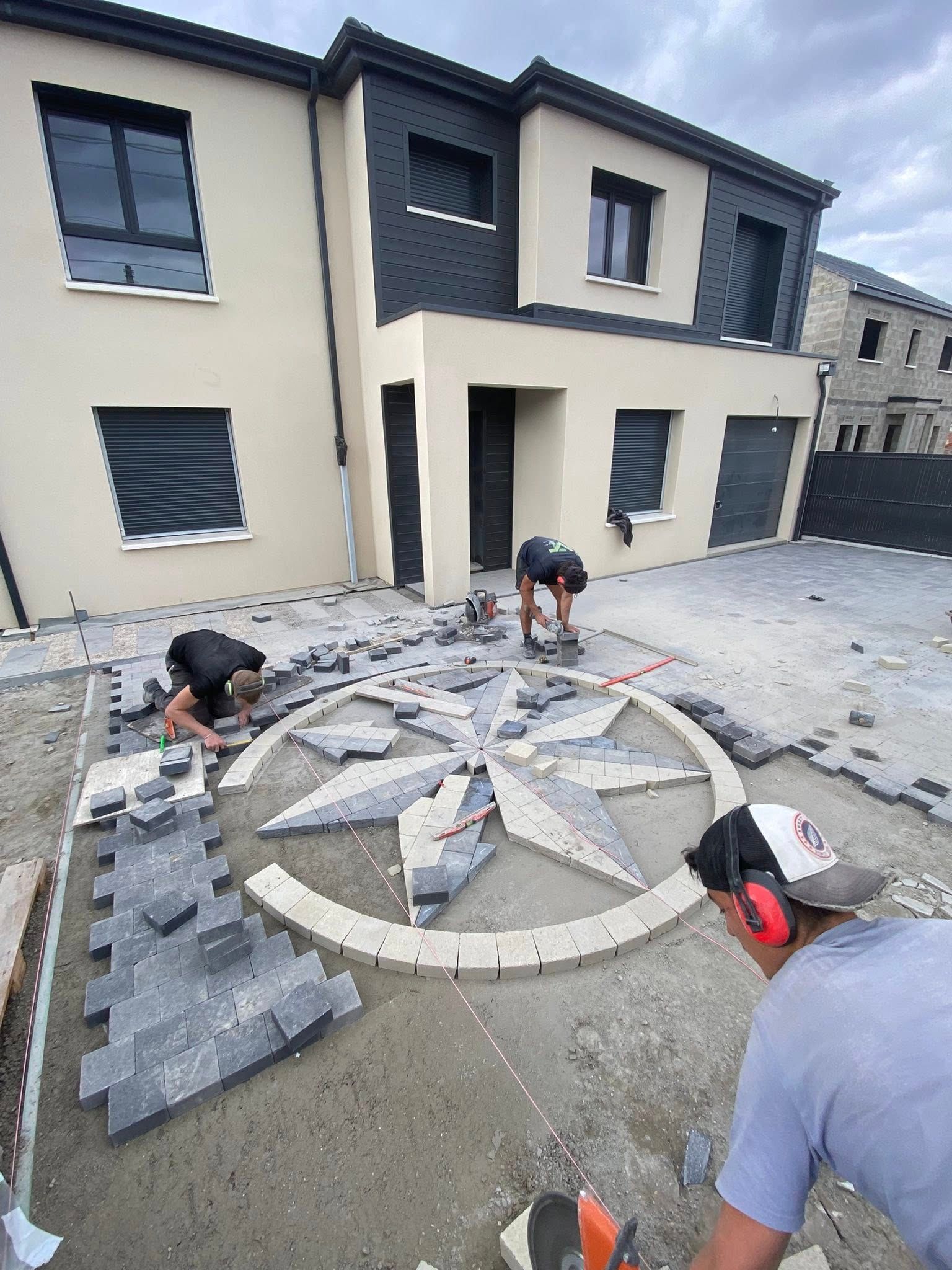 Deux ouvriers installent un motif géométrique de boussole en pierre dans une allée devant une maison moderne.