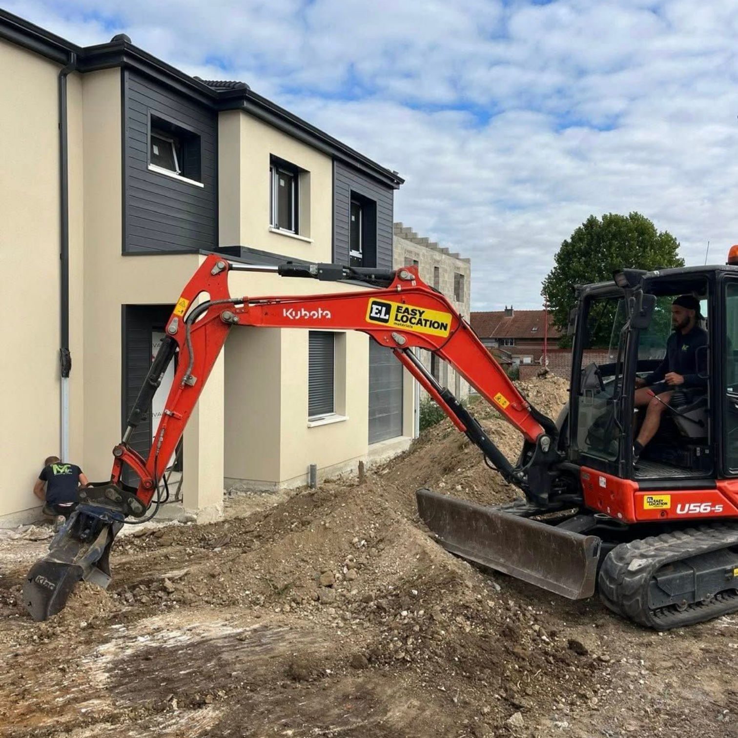 Une pelleteuse rouge creuse une tranchée à côté d'une maison beige moderne, une personne étant accroupie près des fondations.