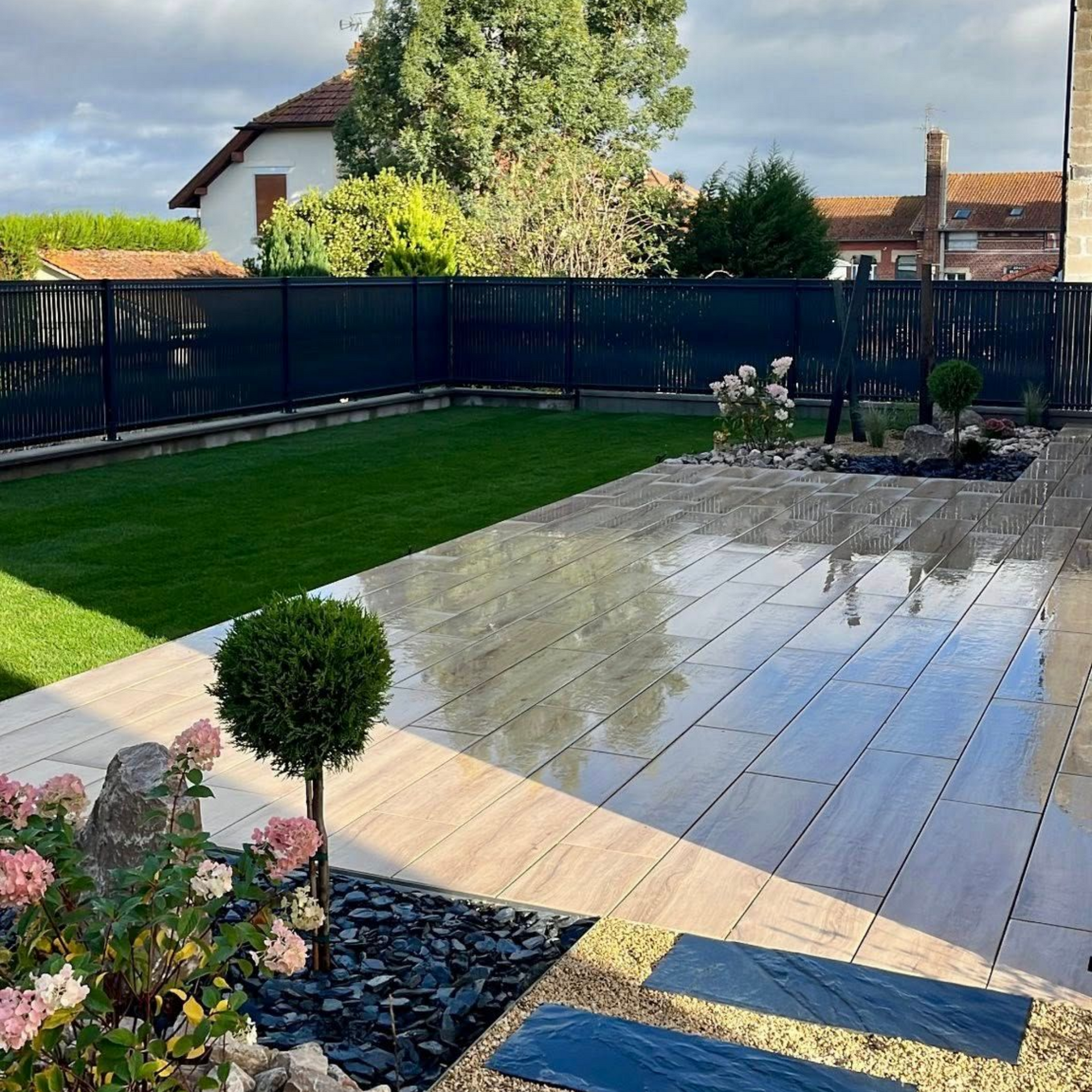 Un jardin avec une terrasse rectangulaire imitation bois, une pelouse, des rosiers dans des parterres d'ardoise et une clôture sombre pour préserver l'intimité.