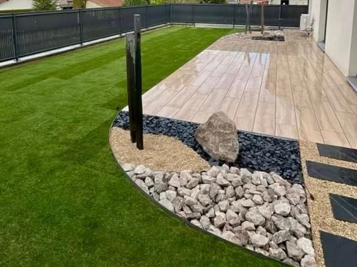 Une scène de jardin avec de la pelouse verte, une terrasse carrelée, du gravier décoratif, des pierres noires et un point d'eau.