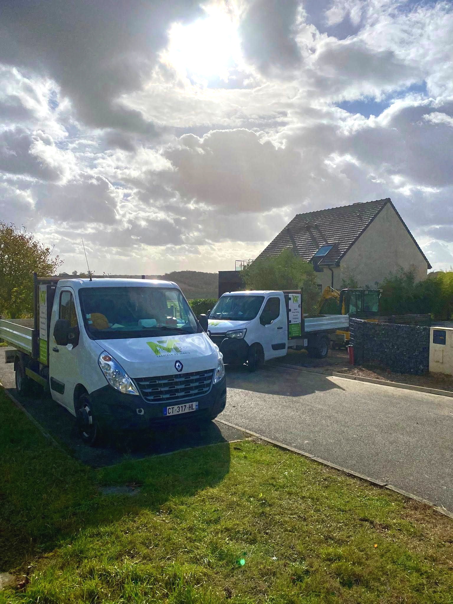 Deux camions utilitaires blancs stationnés en bord de route devant une maison d'habitation, sous un ciel nuageux et ensoleillé.