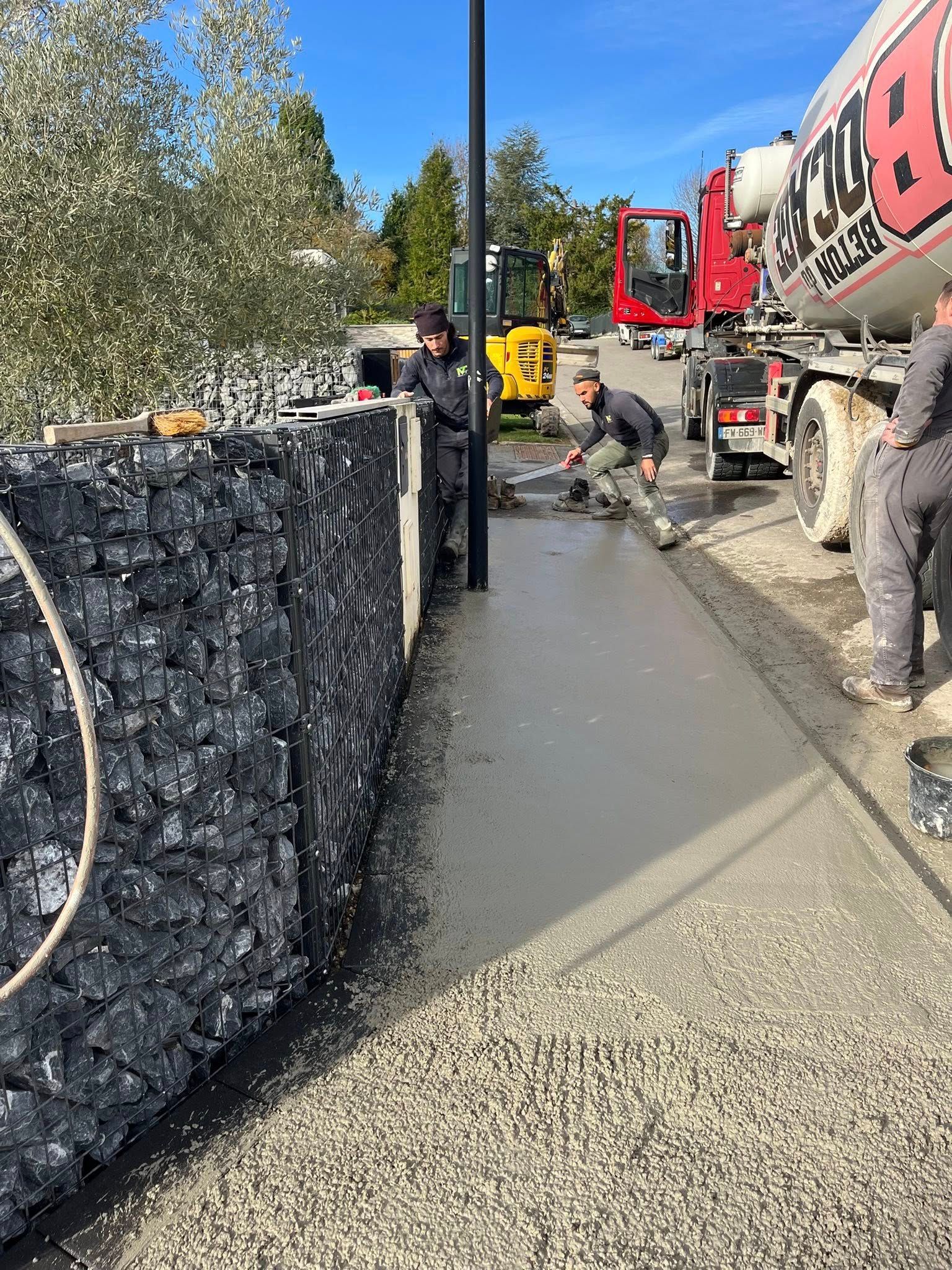 Des ouvriers du bâtiment coulent du béton frais pour un trottoir à côté d'un mur de gabions rempli de pierres et d'un camion-malaxeur à béton stationné.
