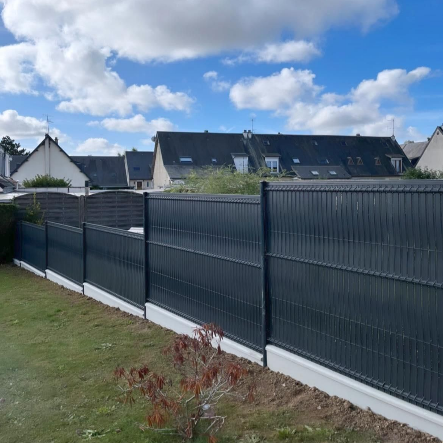Une haute clôture d'intimité gris foncé, composée de lattes verticales, repose sur une base en béton dans une cour sous un ciel bleu clair et nuageux.