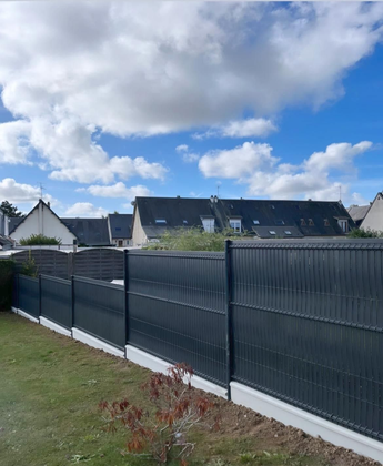 Une clôture d'intimité gris foncé à claire-voie, reposant sur une base en béton, s'étend sur une pelouse sous un ciel bleu nuageux.
