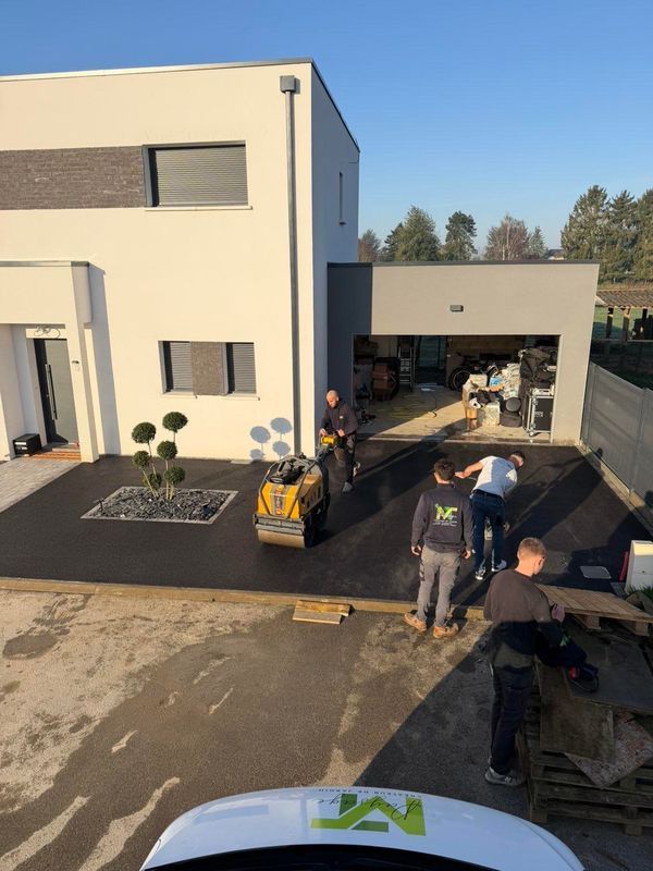 Des ouvriers utilisent un rouleau compresseur pour paver une nouvelle allée en asphalte devant une maison moderne à deux étages.
