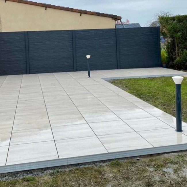 Une terrasse carrelée de couleur claire jouxte une pelouse verte, agrémentée de deux bornes lumineuses et d'une clôture d'intimité gris foncé.