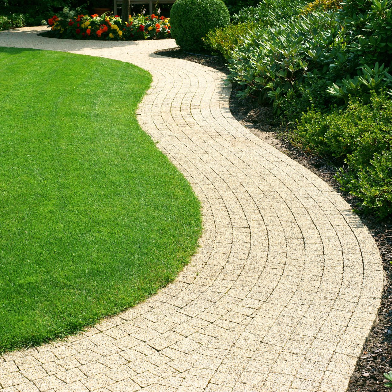 Un chemin pavé clair et sinueux serpente à travers une pelouse verdoyante luxuriante, bordée de plantes et de fleurs de jardin.