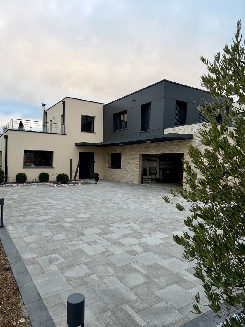 Une maison moderne à deux étages avec un garage revêtu de pierre, un bardage gris foncé, des murs clairs et une allée pavée.