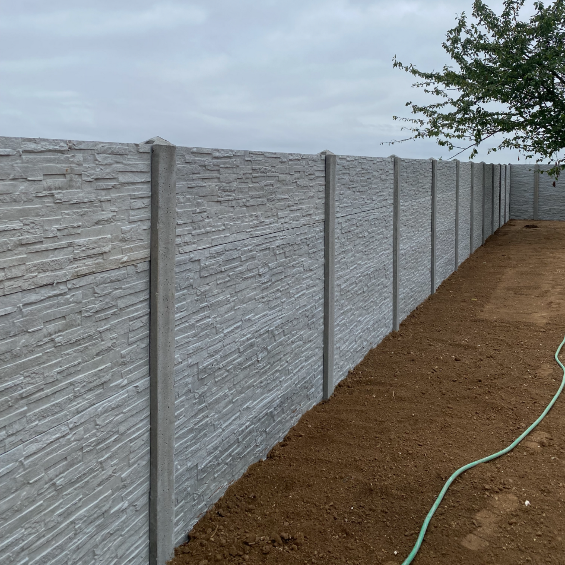 Clôture en béton gris avec des poteaux verticaux le long d'une cour en terre battue, avec un tuyau d'arrosage vert au sol.