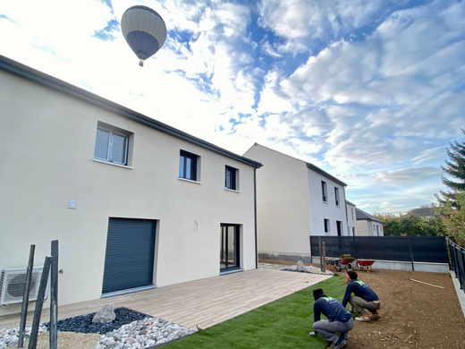 Deux ouvriers posent du gazon dans un jardin à côté d'une nouvelle maison à deux étages, avec une montgolfière dans le ciel bleu nuageux.