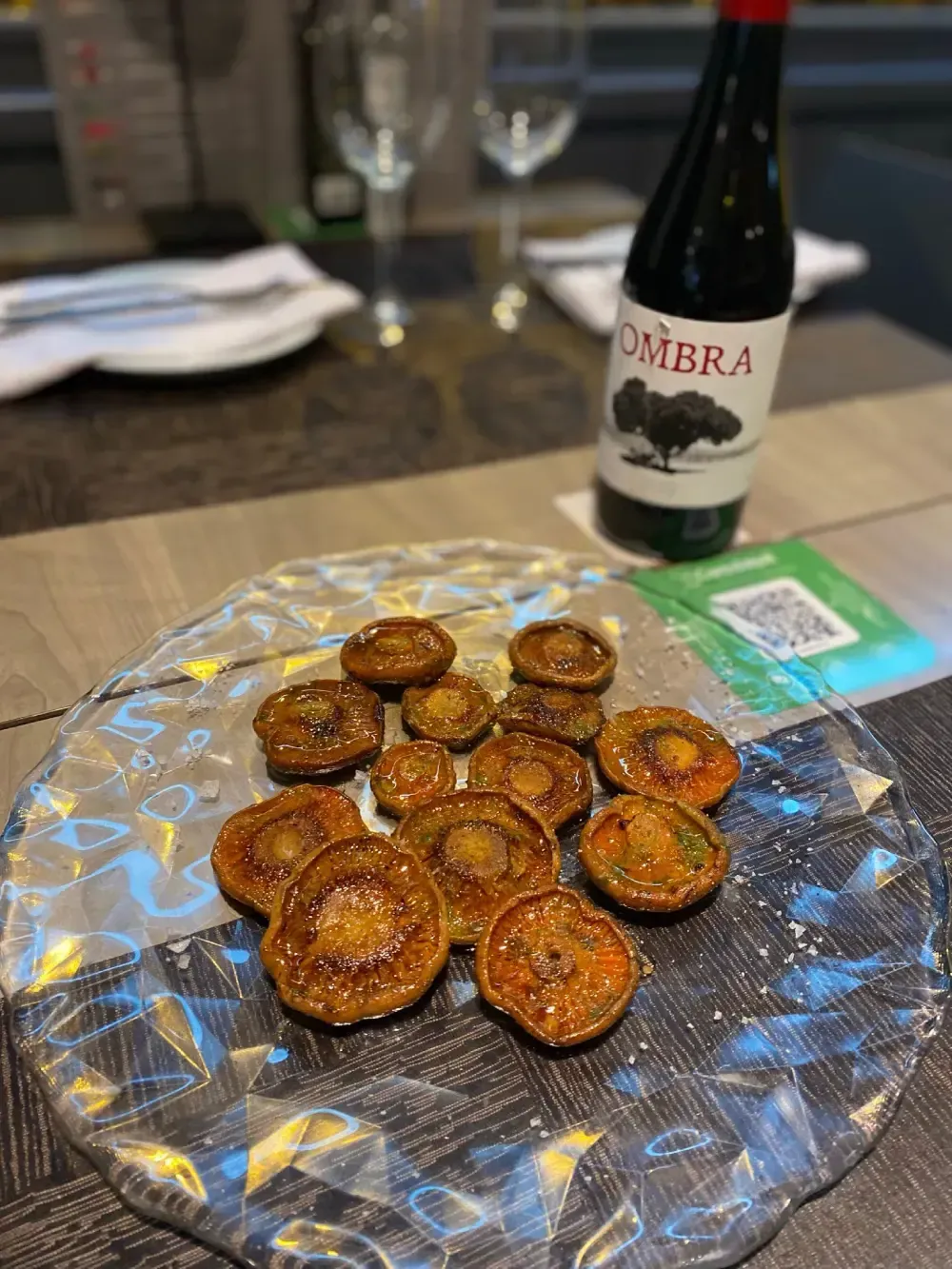 Plato de champiñones cocidos sobre una mesa, con una botella de vino al fondo.