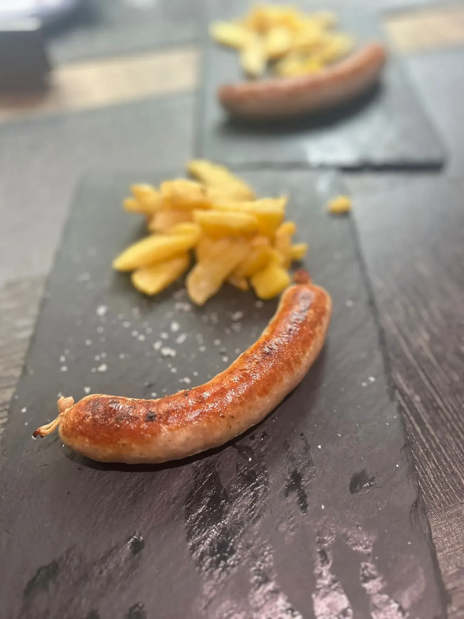 Salchicha a la parrilla con patatas fritas sobre una pizarra negra, con sal. Otro plato similar al fondo.