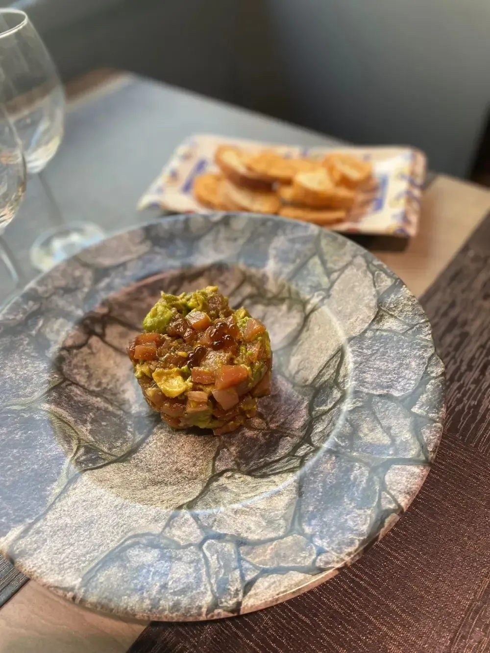 Aperitivo de aguacate y tomate en un plato estampado con pan tostado de fondo.