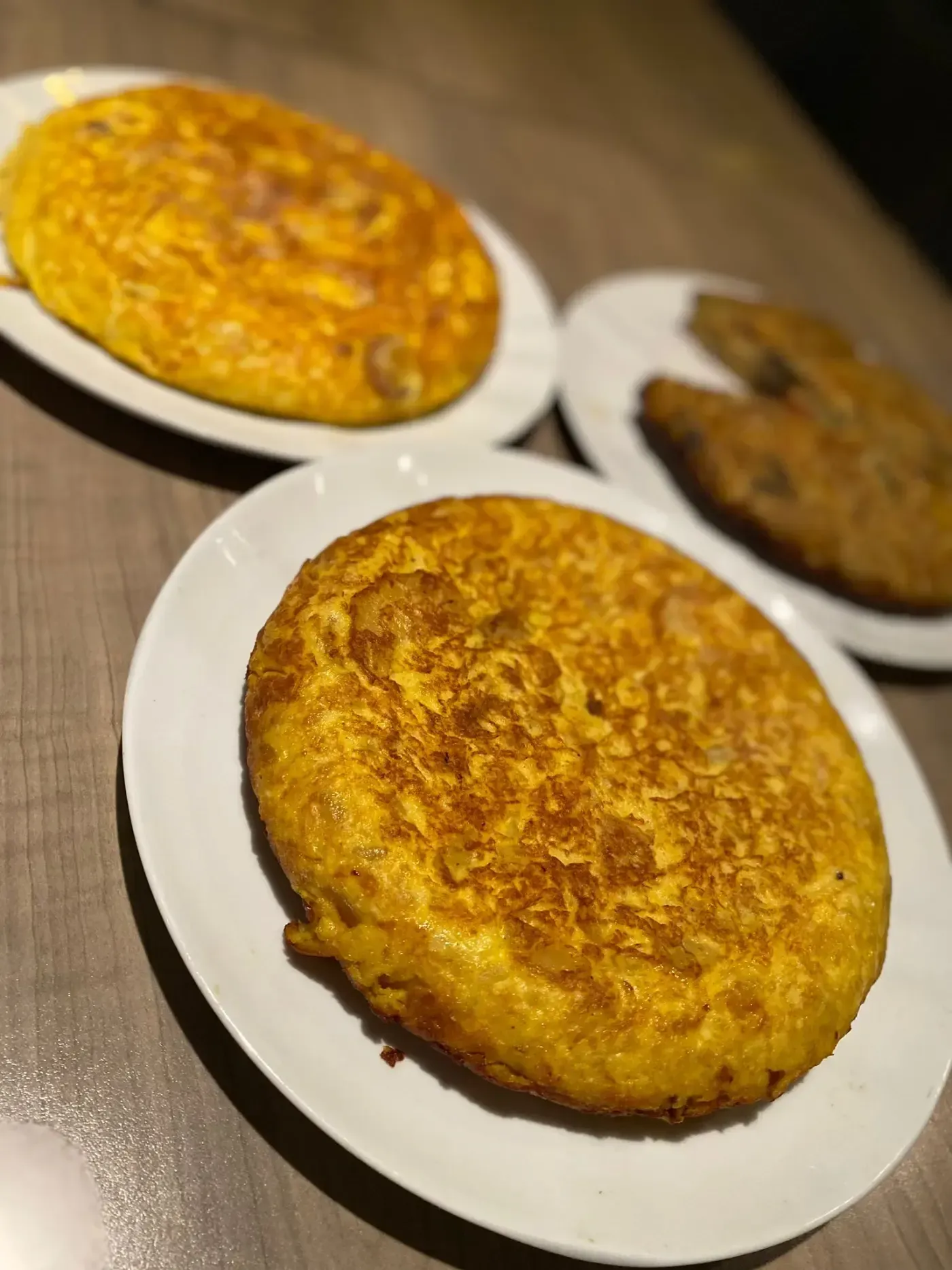 Tres tortillas españolas en platos blancos, de color amarillo dorado, colocados sobre una mesa de madera.