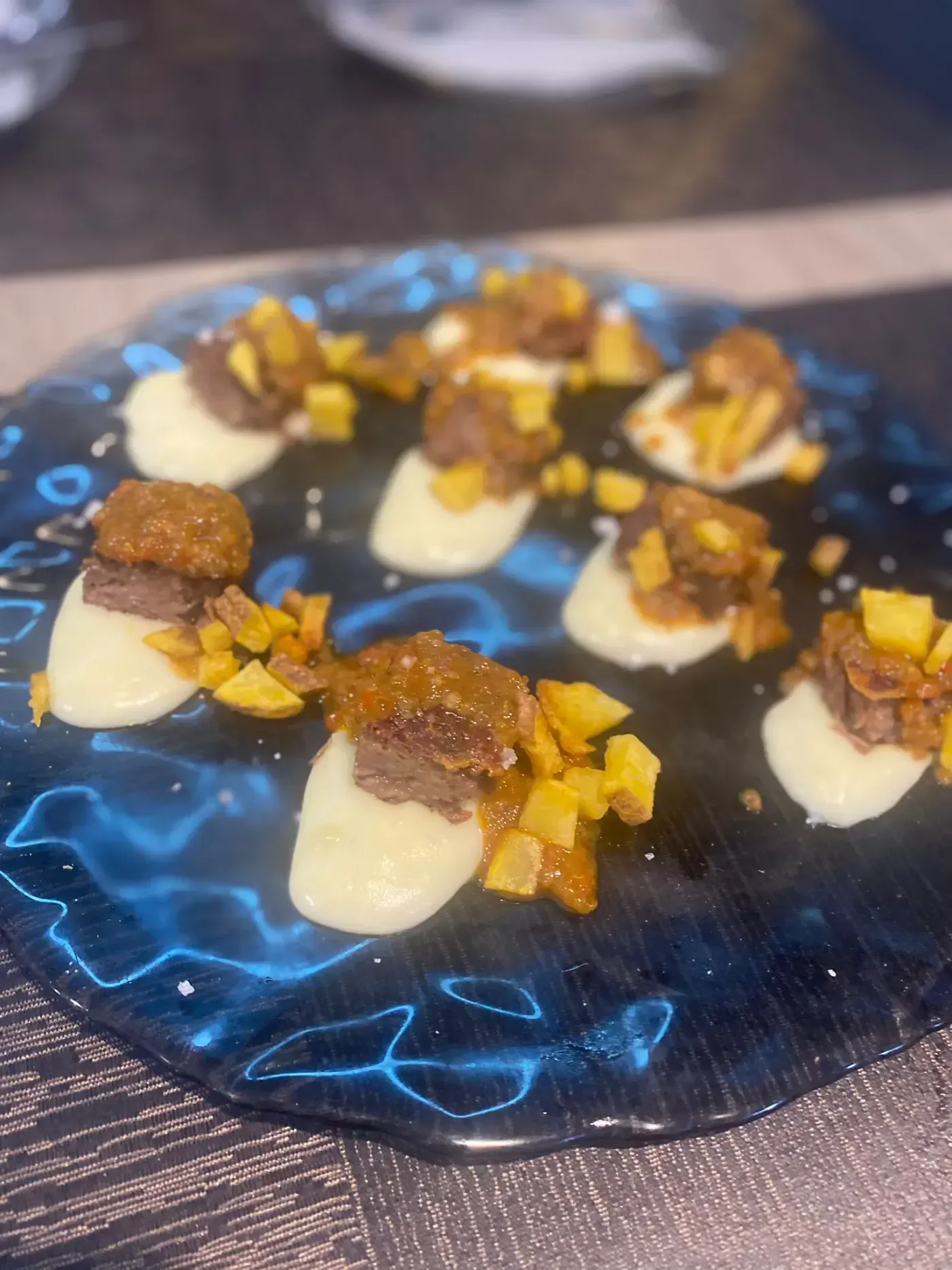 Aperitivos en un plato de cristal azul: puré blanco, patatas cortadas en cubitos y cubierto con carne oscura y salsa.
