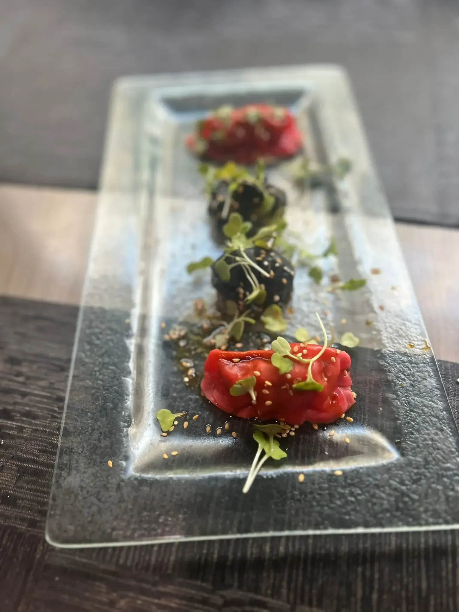 Cuatro arreglos de comida coloridos en un plato de vidrio rectangular, adornados con brotes y semillas de sésamo.