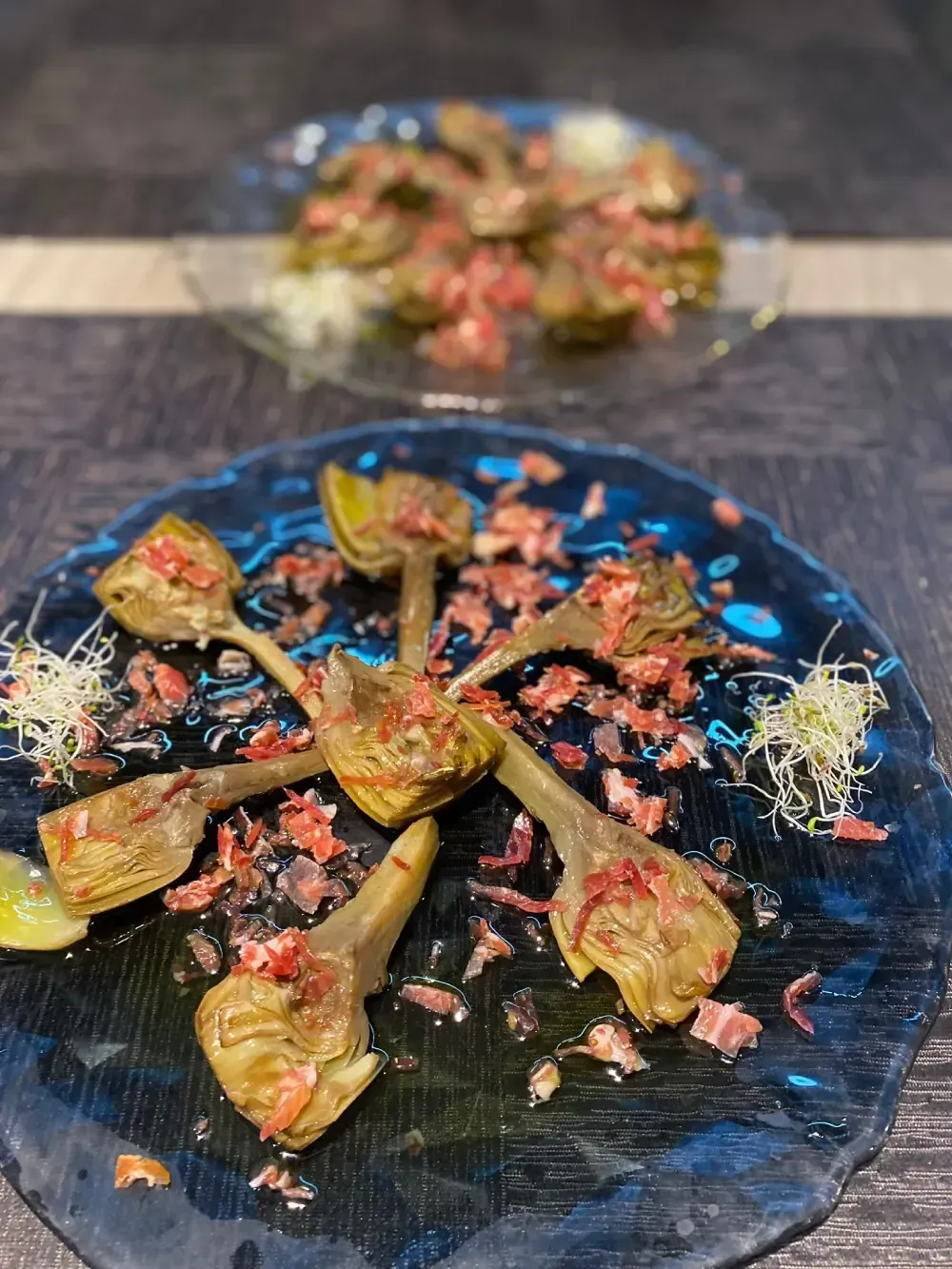 Corazones de alcachofa con jamón en un plato oscuro, otro plato de comida similar al fondo.