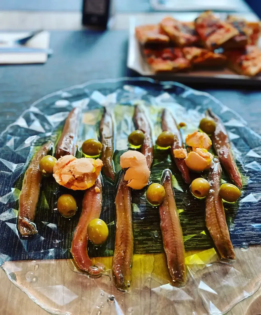 Anchoas en aceite de oliva, adornadas con aceitunas y posiblemente pan rallado en un plato transparente.
