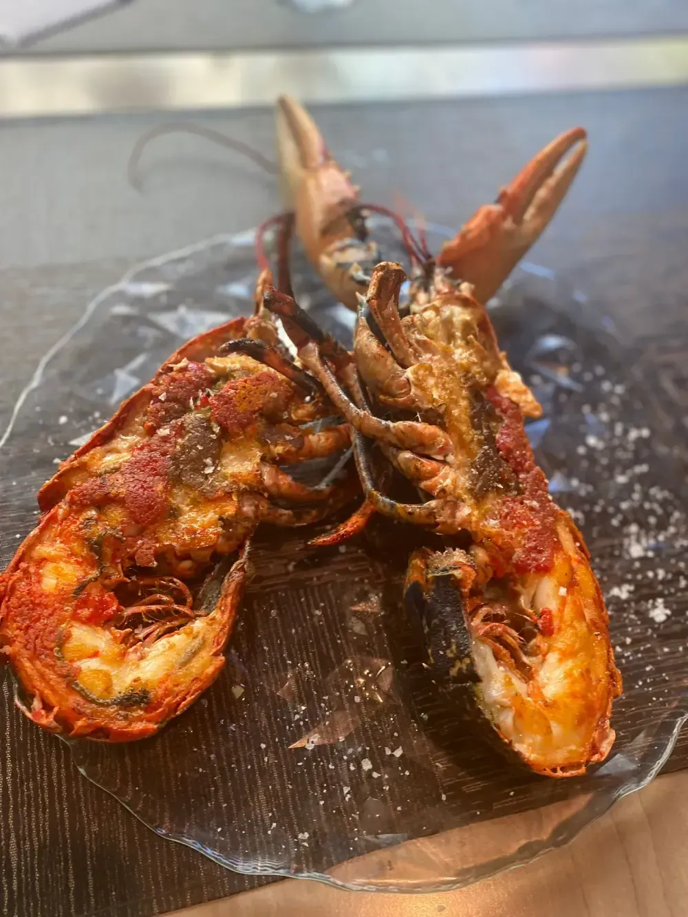 Dos mitades de langosta a la parrilla en un plato transparente, con salsa roja y sal espolvoreada.