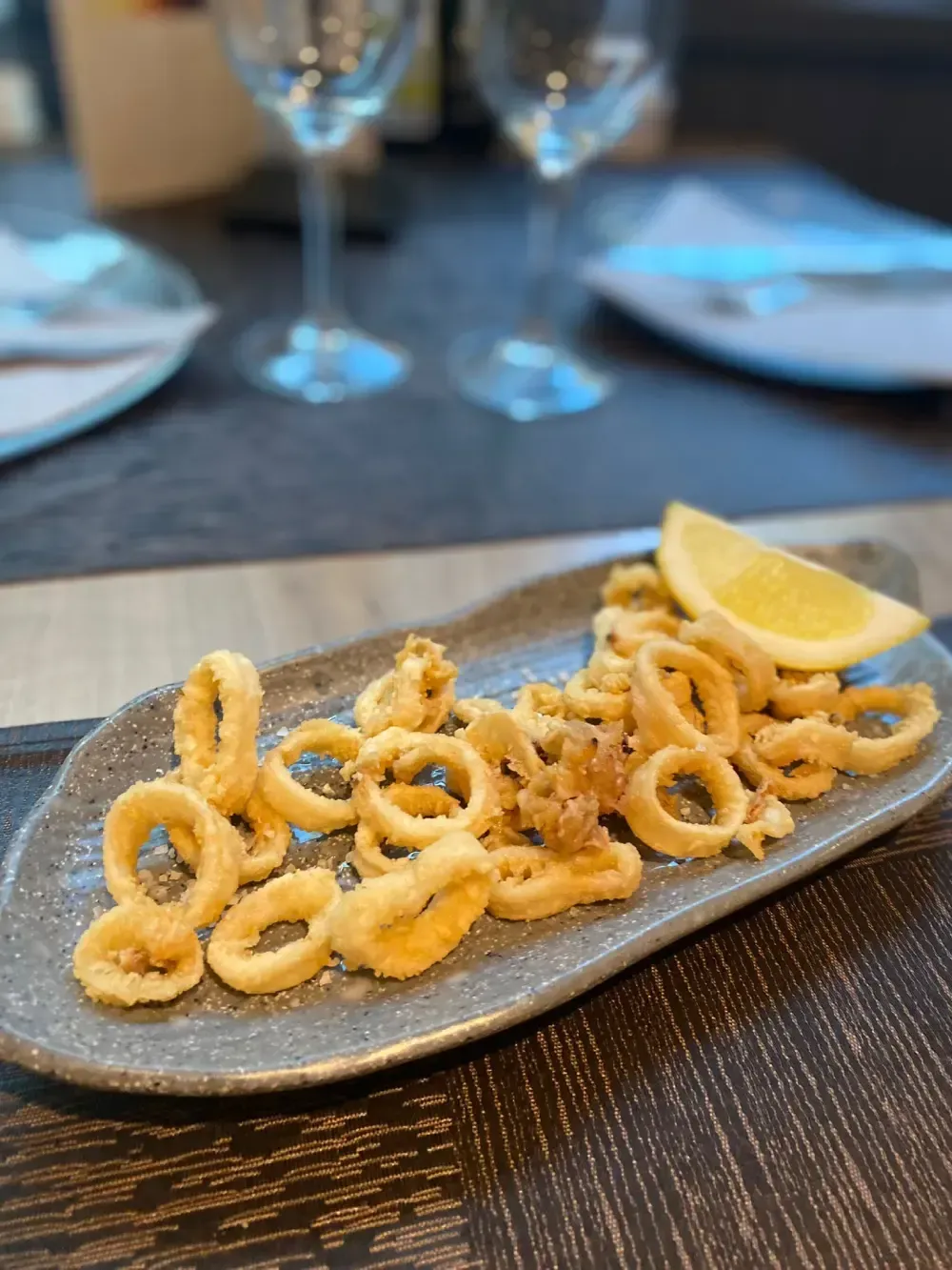 Anillos de calamares fritos en un plato gris con una rodaja de limón, colocado sobre la mesa de un restaurante.