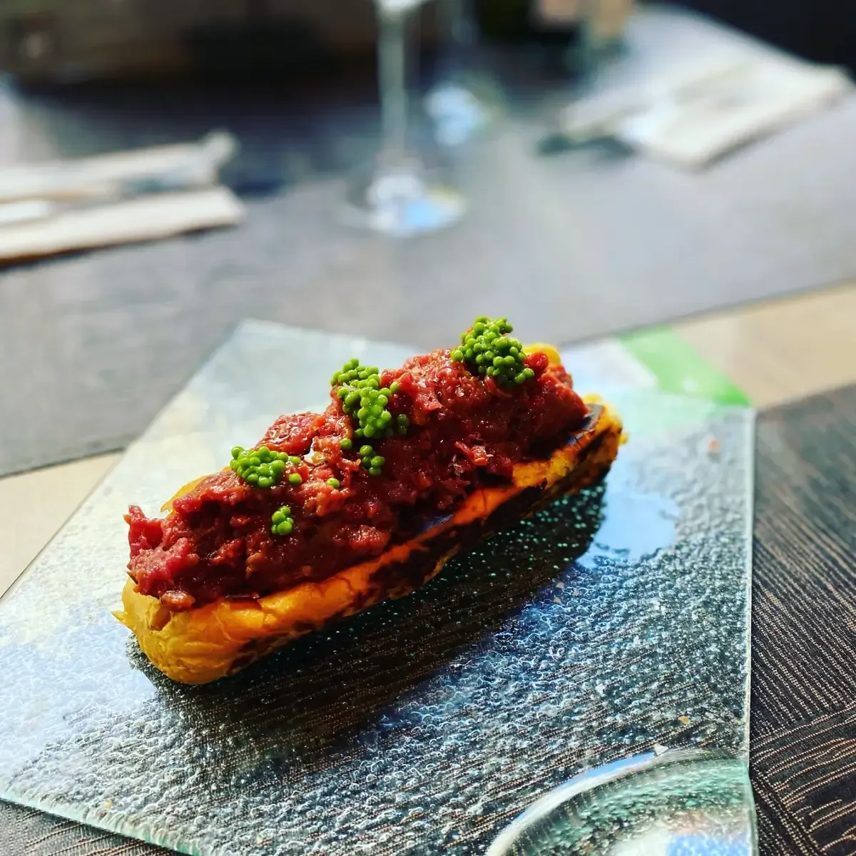 Aperitivo: Una rebanada de pan tostado cubierta con carne roja y hierbas verdes en un plato de vidrio.