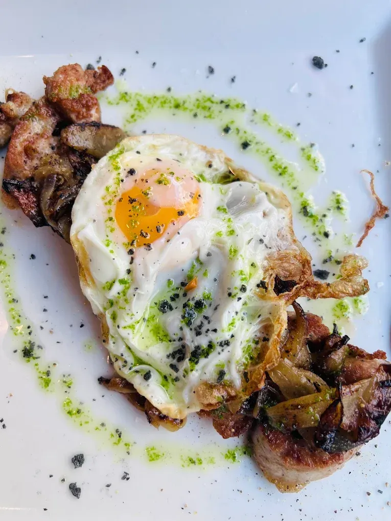 Huevo frito con yema líquida en un plato con tocino y un chorrito de salsa verde.