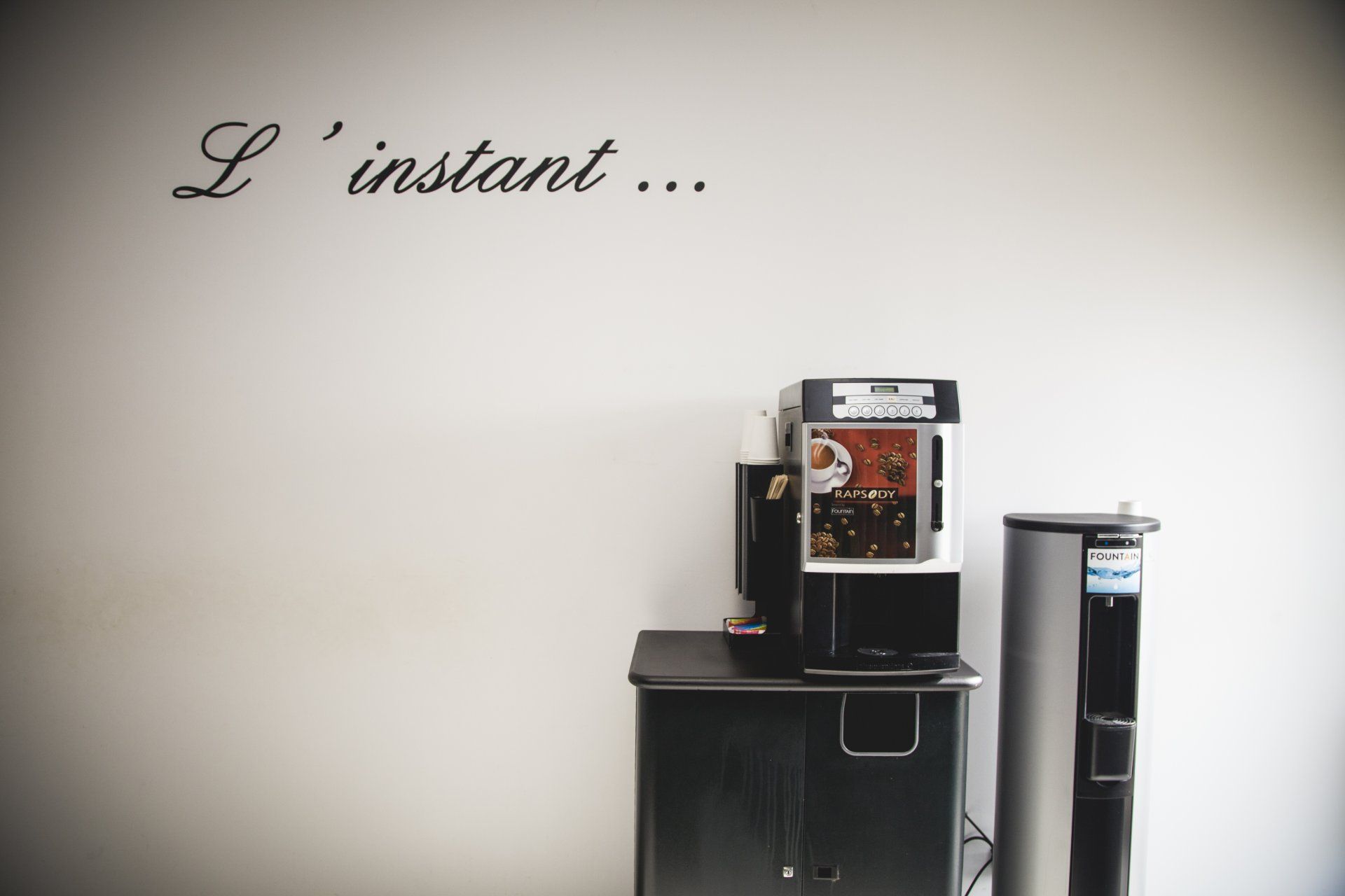 Machine à café et fontaine à eau contre un mur blanc avec l'inscription « L'instant... » en lettres cursives.