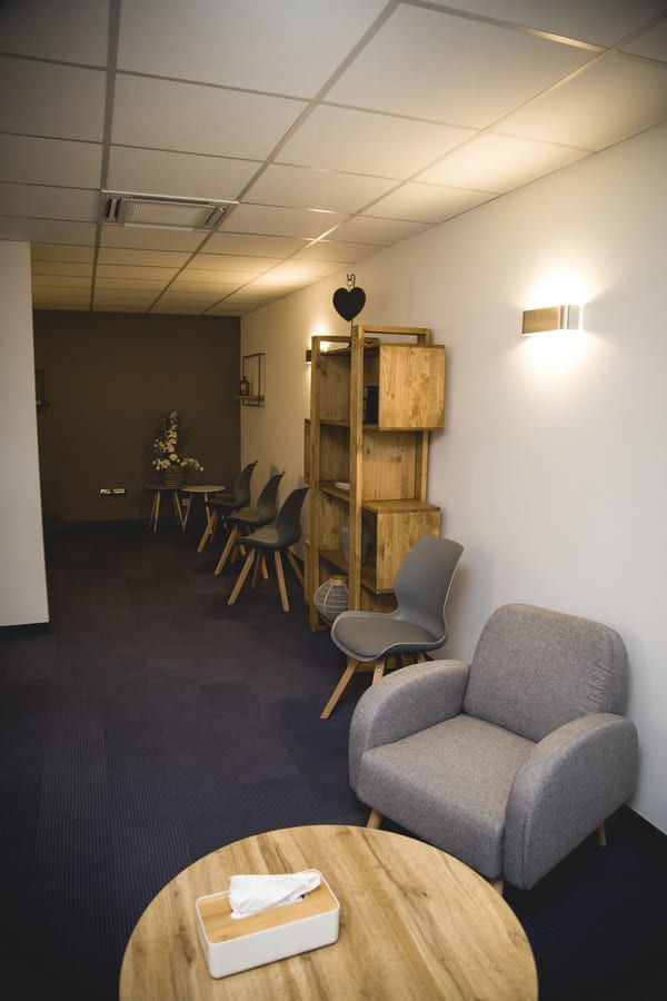 Salle d'attente avec chaises grises, étagères en bois, table ronde et moquette bleue.