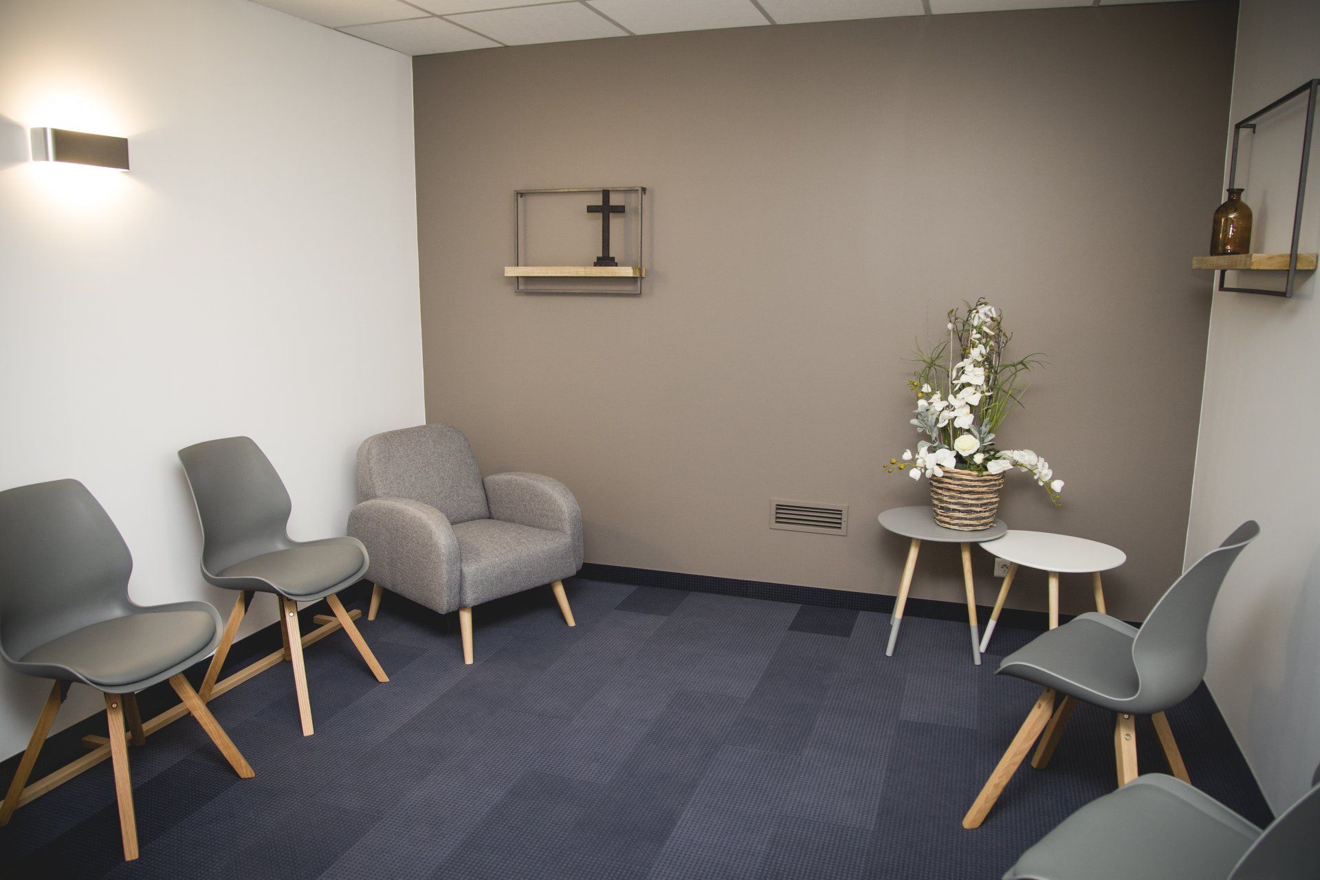 Salle d'attente avec chaises grises, un fauteuil, des petites tables et une composition florale.