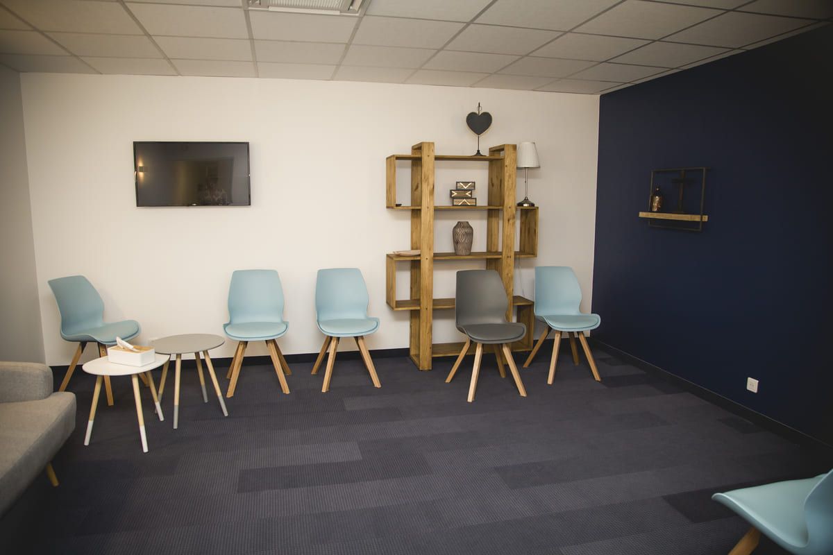 Salle d'attente avec chaises bleues, tables basses et étagère en bois sur fond de murs bleus et blancs.