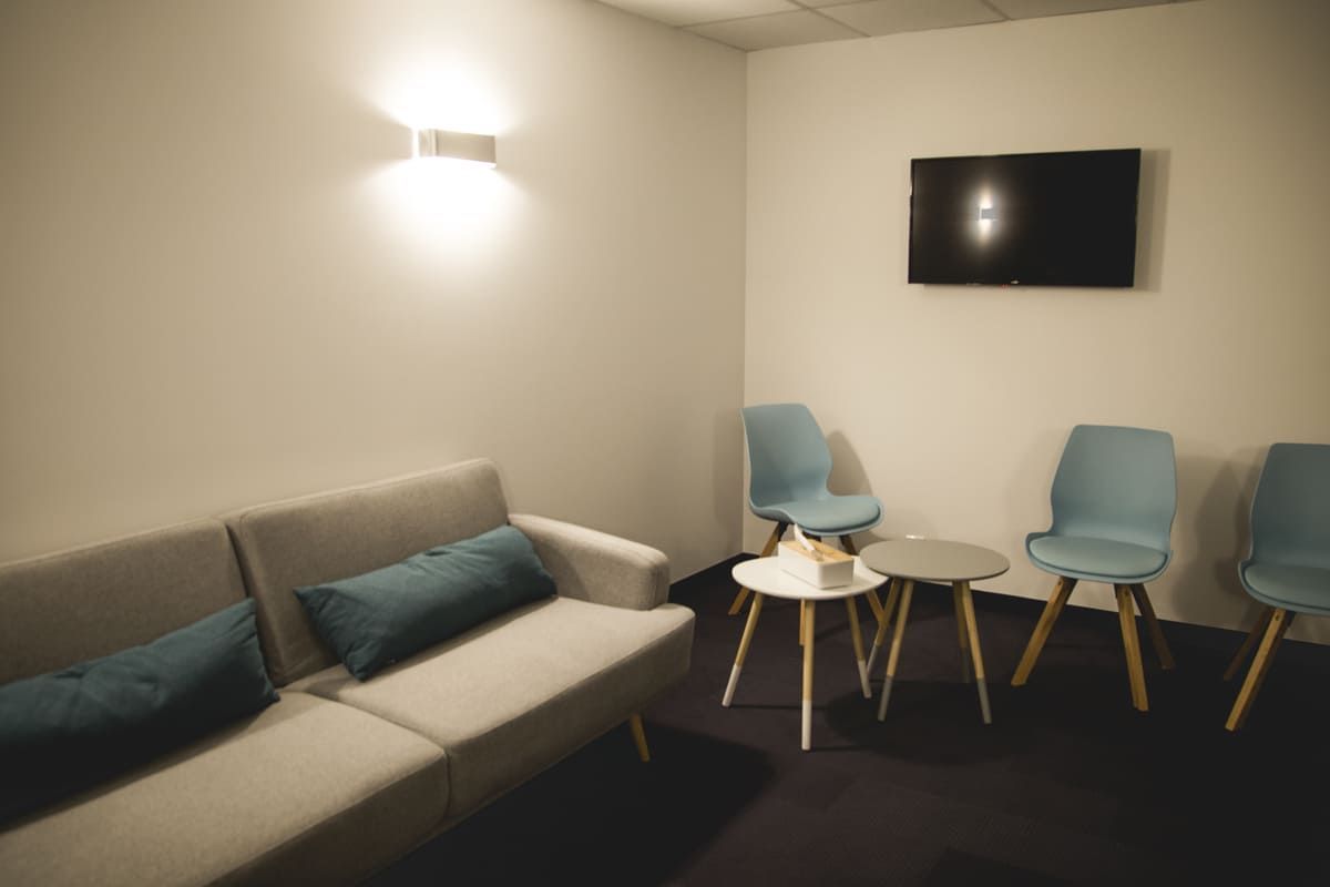 Salle d'attente avec canapé gris, chaises turquoise, petites tables et télévision murale.