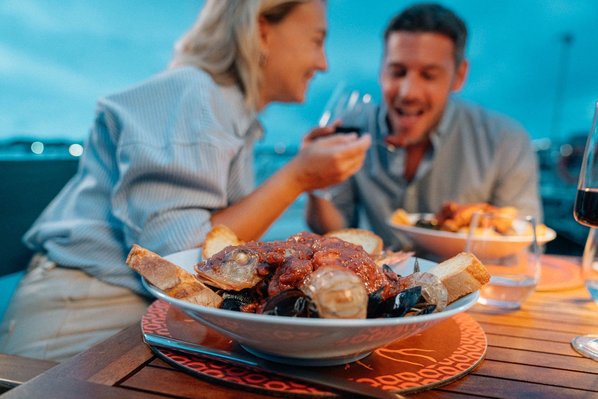 A man and a woman are sitting at a table eating food and drinking wine.