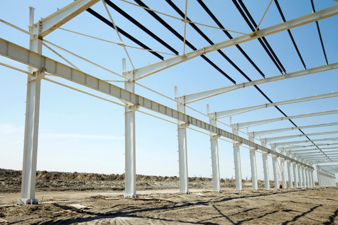 Weißes Stahlgerüst eines im Bau befindlichen Gebäudes vor blauem Himmel und braunem Erdboden.