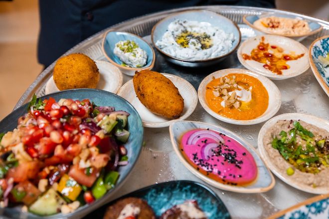 Innenansicht eines schwach beleuchteten Restaurants mit Bar, Hockern, Tischen und verschwommenen Gestalten von Personal und Gästen.