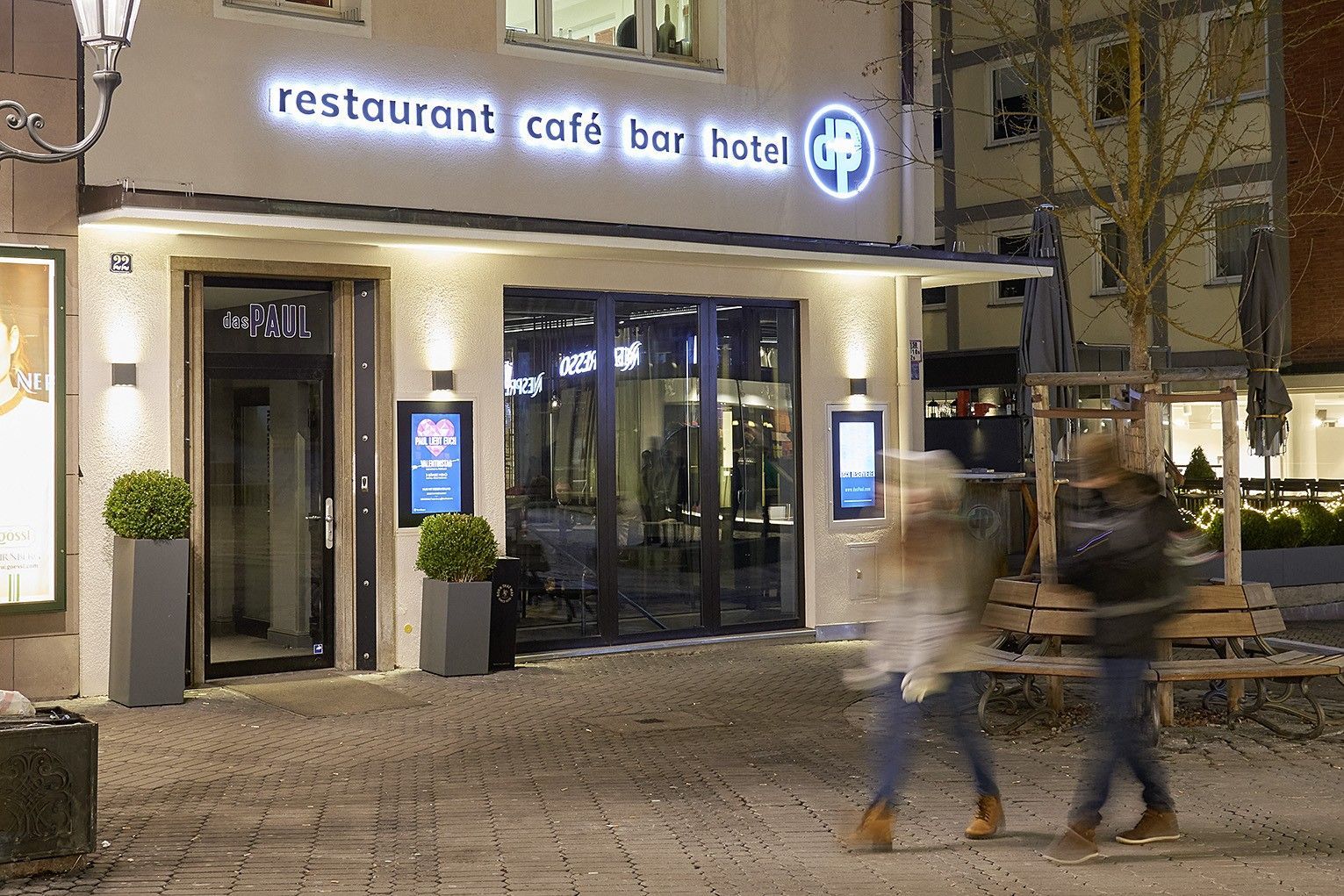 Menschen gehen vor einem Restaurant, auf dem Restaurant Café Bar Hotel steht