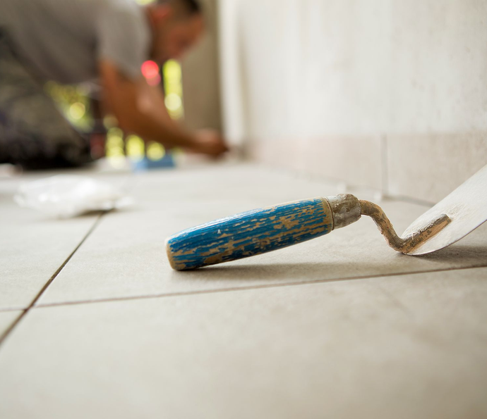 Truelle sur un sol carrelé, avec une personne floue posant des carreaux en arrière-plan.