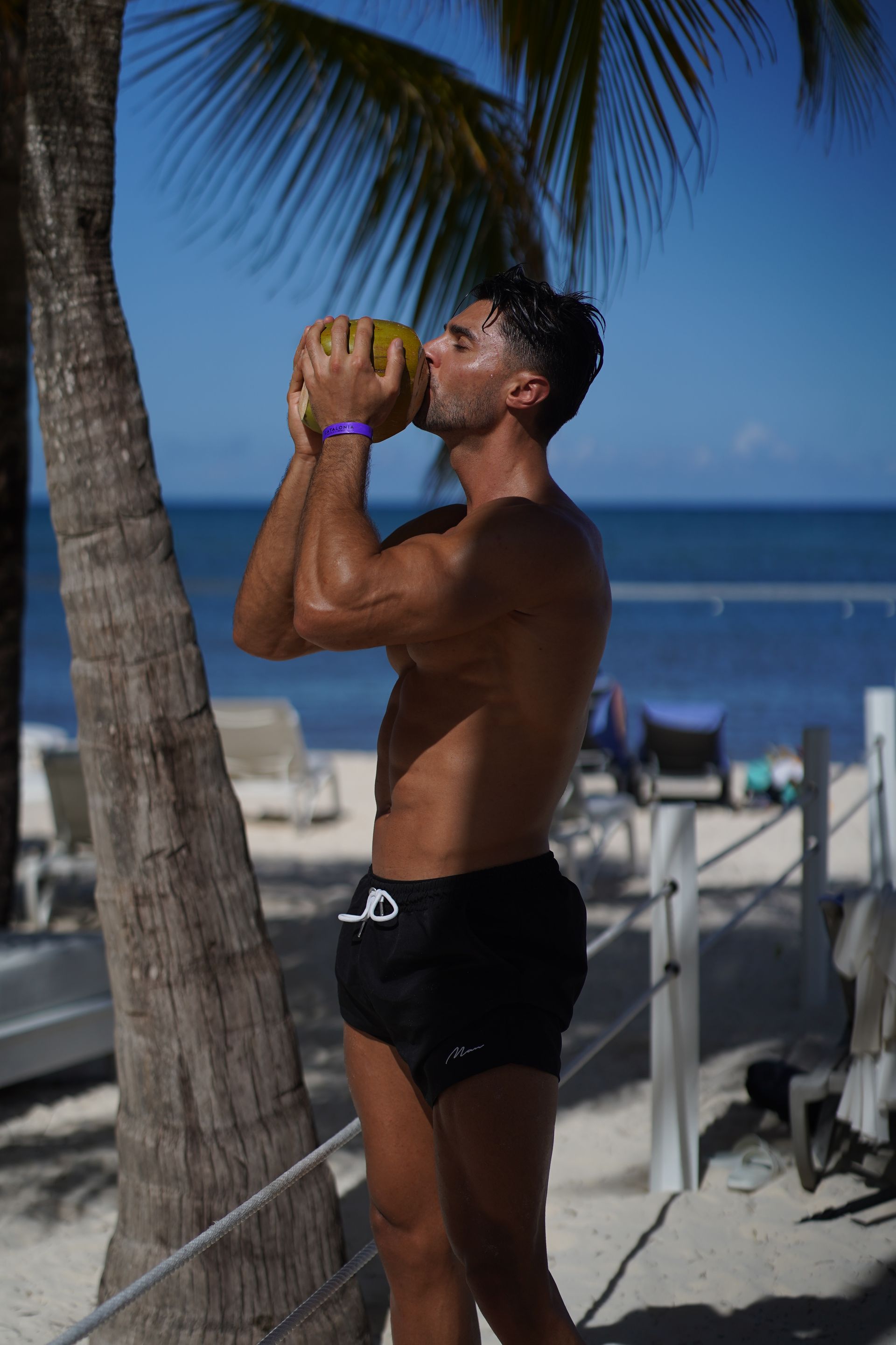 Ein Mann ohne Hemd trinkt am Strand aus einer Kokosnuss.