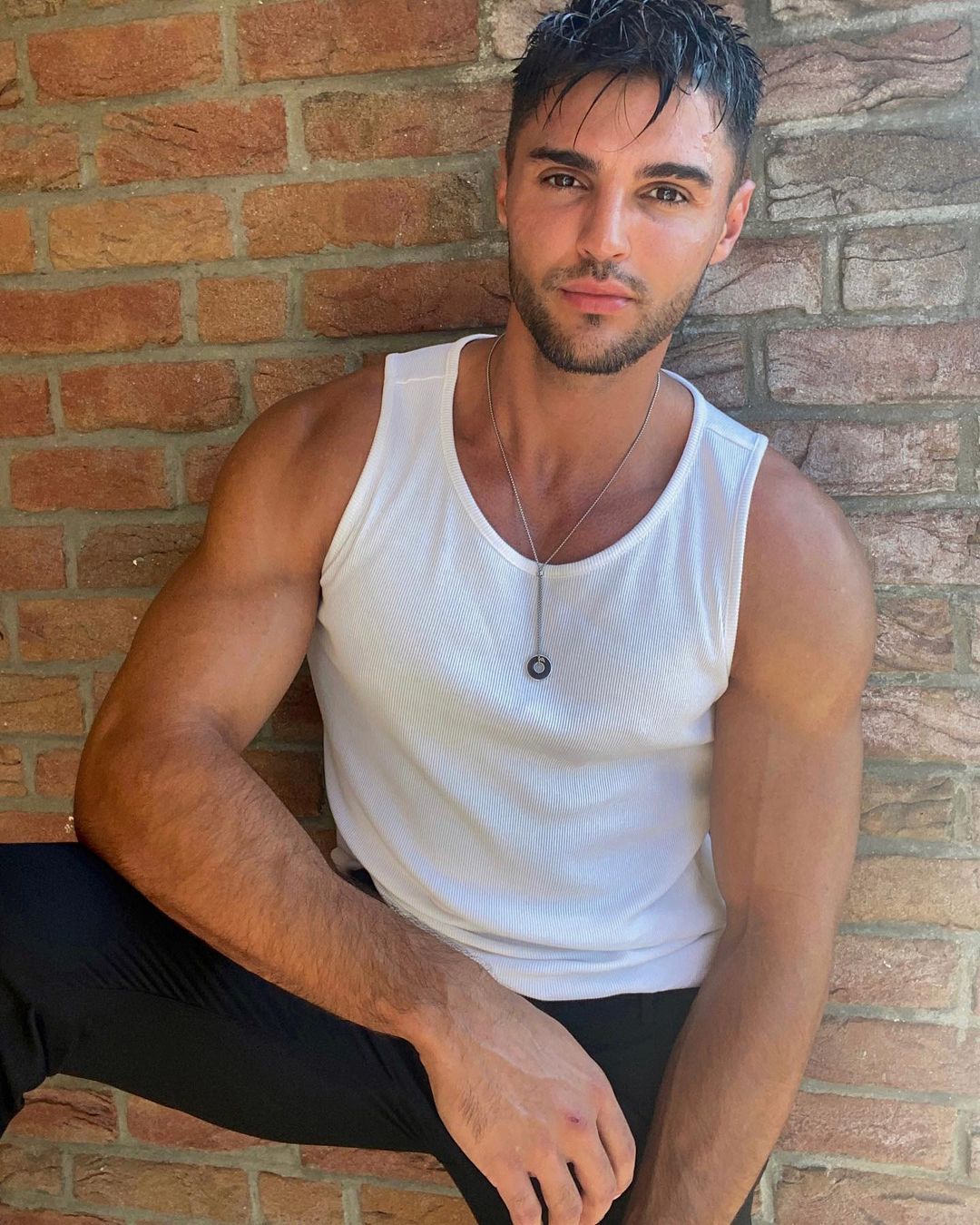 Ein Mann in einem weißen Tank Top sitzt vor einer Backsteinmauer.