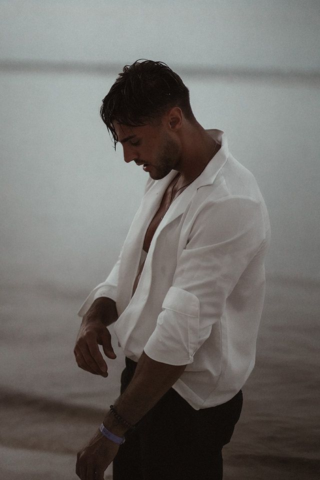 Ein Mann im weißen Hemd steht am Strand und schaut auf seine Uhr.