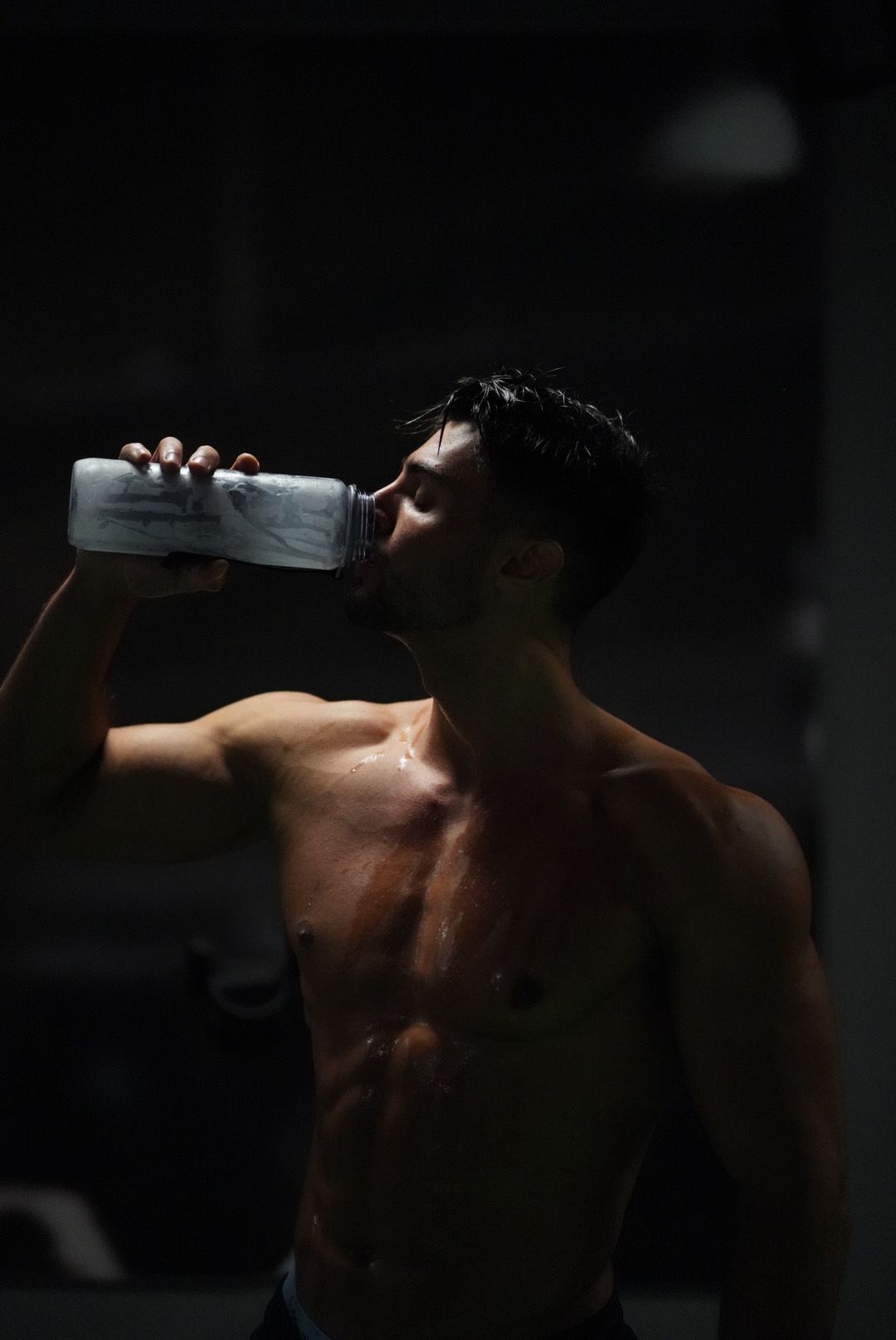 Ein Mann ohne Hemd trinkt in einem dunklen Raum Wasser aus einer Flasche.