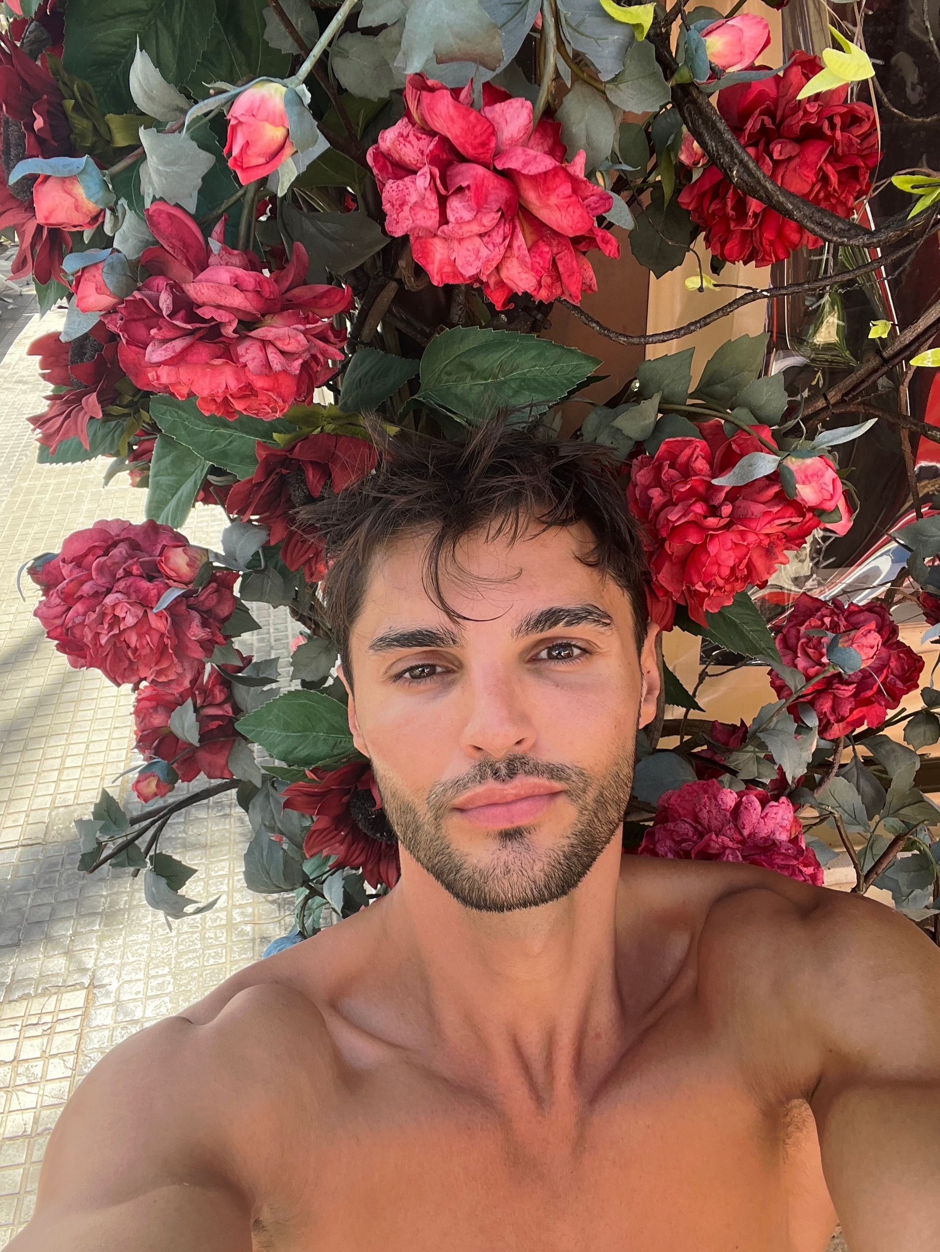 Ein Mann ohne Hemd macht ein Selfie vor einem Baum mit roten Blüten.