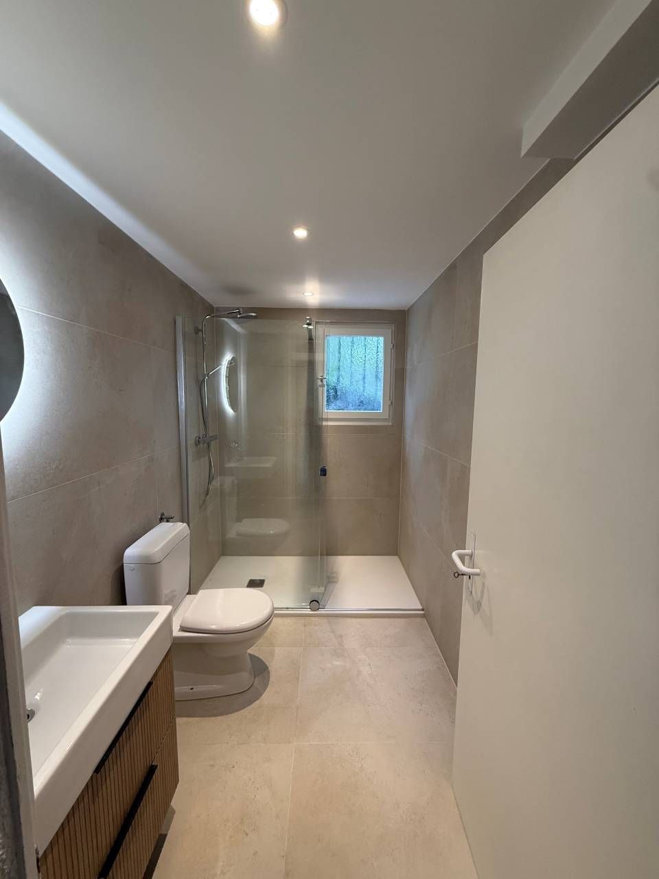 Salle de bains moderne aux tons neutres, douche, toilettes et lavabo. Porte blanche à droite.