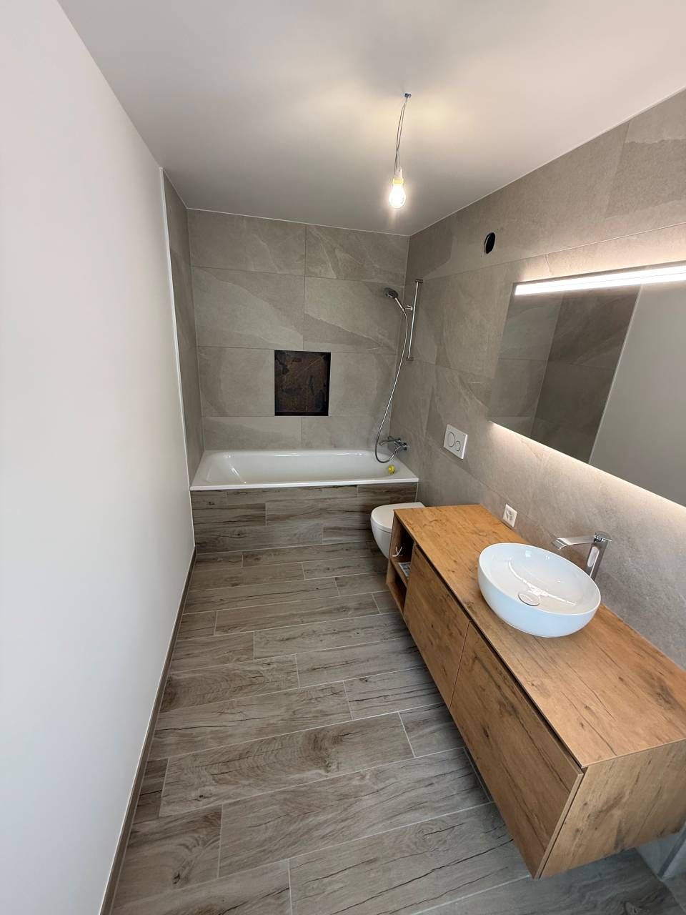 Salle de bains moderne avec sol imitation bois, murs carrelés gris, lavabo et baignoire.