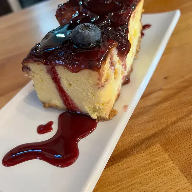 Una rebanada de tarta de queso con un arándano encima en un plato blanco