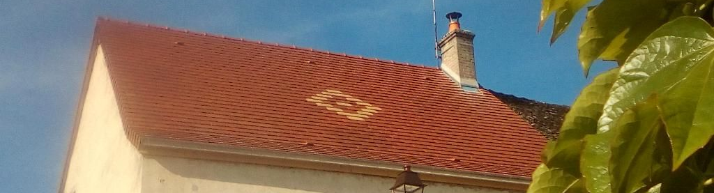 Toit de tuiles rouges d'un bâtiment avec cheminée se détachant sur un ciel bleu, quelques feuilles vertes au premier plan.