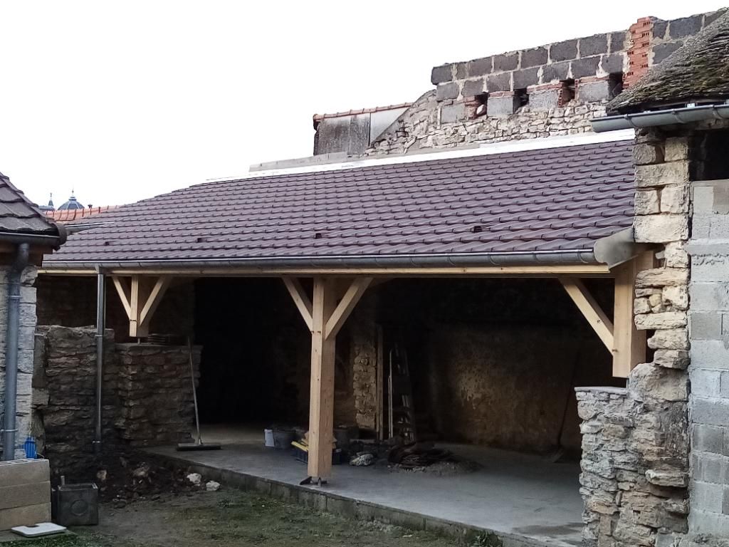 Abri de voiture avec toit de tuiles brunes, soutenu par des poutres en bois, construit entre des murs en pierre.