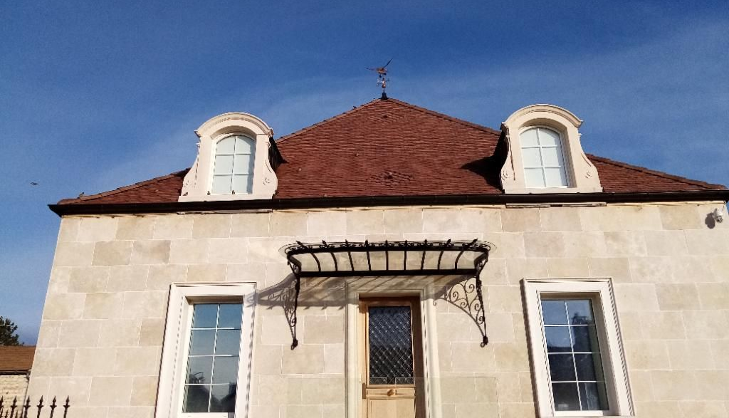 Maison en pierre au toit de tuiles rouges, avec lucarnes et auvent en métal noir, se détachant sur un ciel bleu.