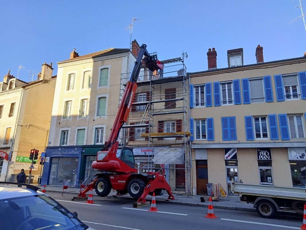 Une grue de chantier rouge soulève des poutres près de bâtiments aux volets bleus et aux échafaudages.