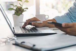 Personne tapant sur un ordinateur portable, tenant son téléphone, un stylo, une tasse de café et une plante sur son bureau.